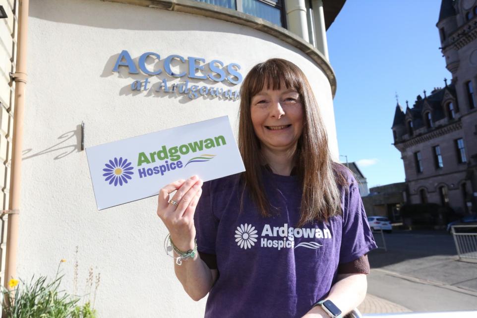 A smiling woman stands outside a building with a sign reading "ACCESS at Ardgowan." She is wearing a purple "Ardgowan Hospice" T-shirt and holding a sign with the Ardgowan Hospice logo, which features a purple daisy and green and blue text. The weather appears sunny, and an old stone building is visible in the background.