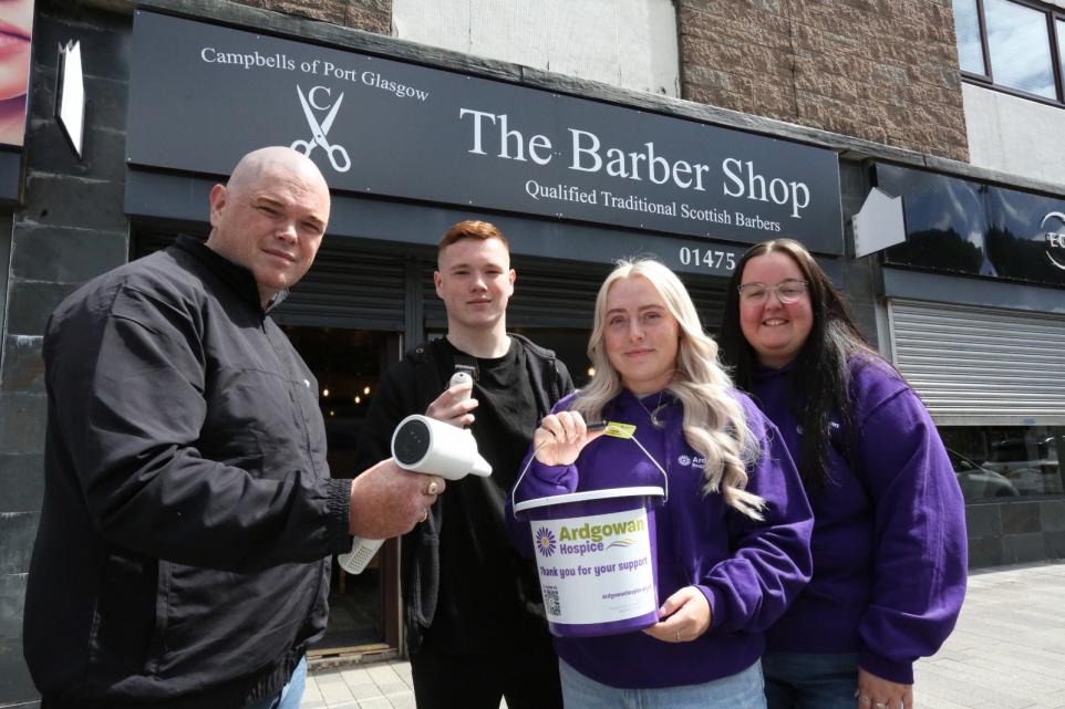 Four people stand outside "The Barber Shop" in Port Glasgow. Two men on the left hold barber tools, while two women on the right wear purple Ardgowan Hospice hoodies. One of the women holds a donation bucket labeled "Ardgowan Hospice – Thank you for your support." All are smiling in support of the hospice.