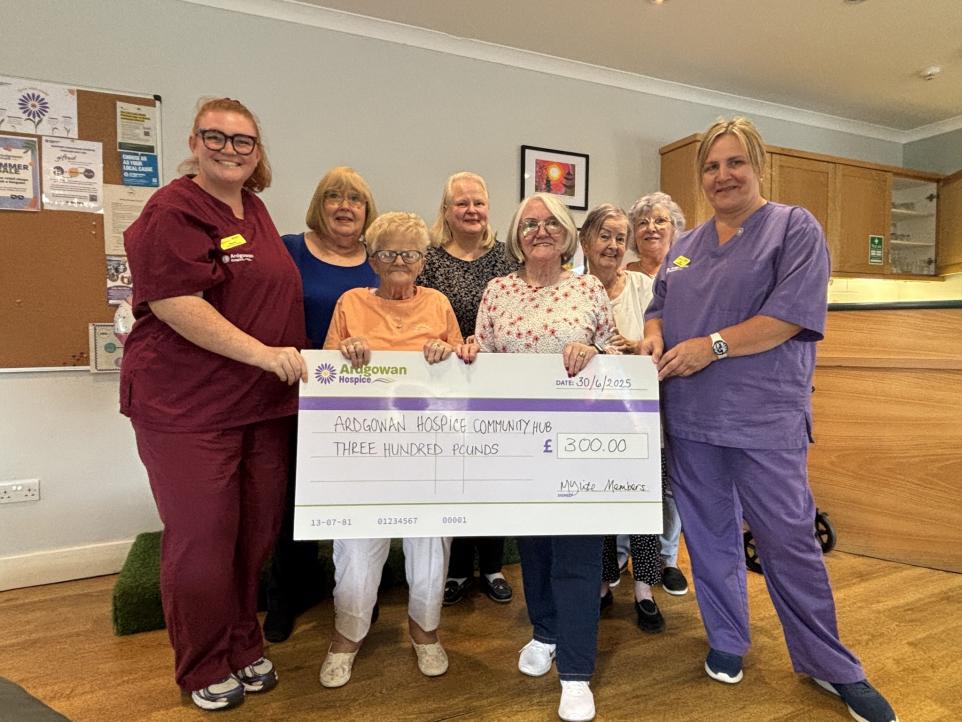 A group of nine women, including two healthcare workers in scrubs, holding a large ceremonial check for £300 made out to Ardgowan Hospice Community Hub. The women are smiling and standing indoors, with a bulletin board and wooden furniture visible in the background.