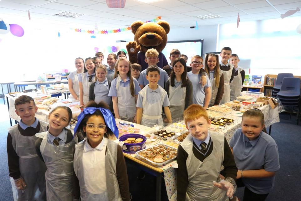A group of children in school uniforms and aprons are gathered around tables filled with baked goods and treats in a decorated classroom. They are smiling and posing for the photo, with a person dressed as a large teddy bear mascot Ace standing behind them. The classroom is decorated with colourful balloons and hanging paper decorations, suggesting a celebration or special event.