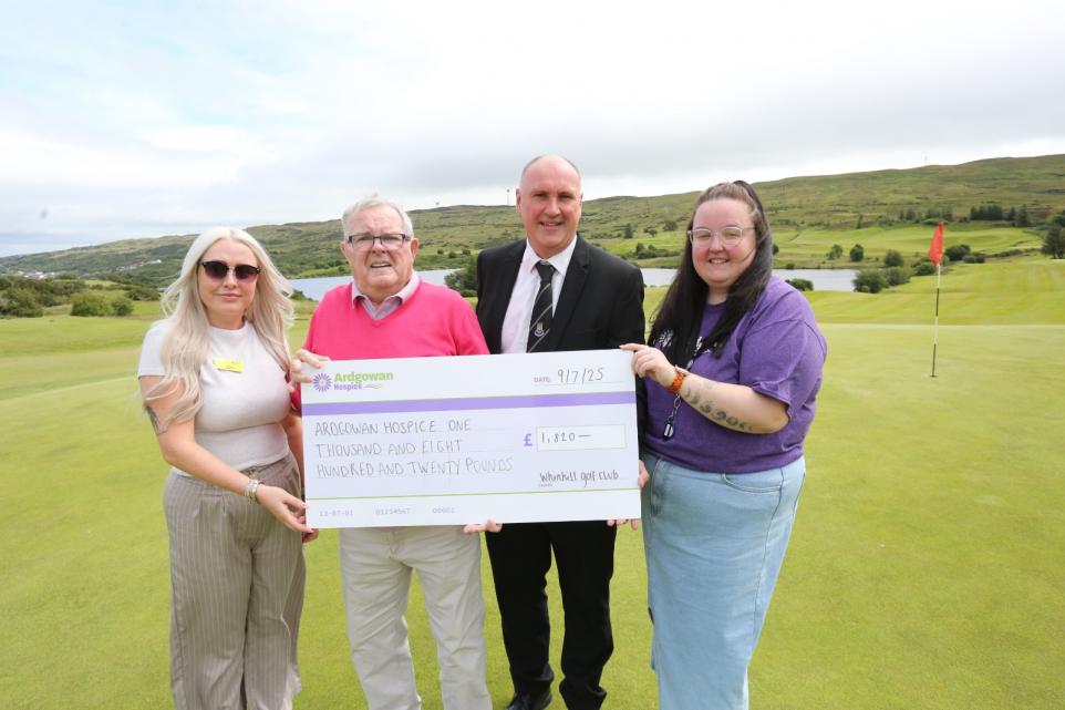 A group of four people standing on a golf course, holding a large cheque made out to Ardgowan Hospice for £1,820. The background features rolling green hills and a cloudy sky.
