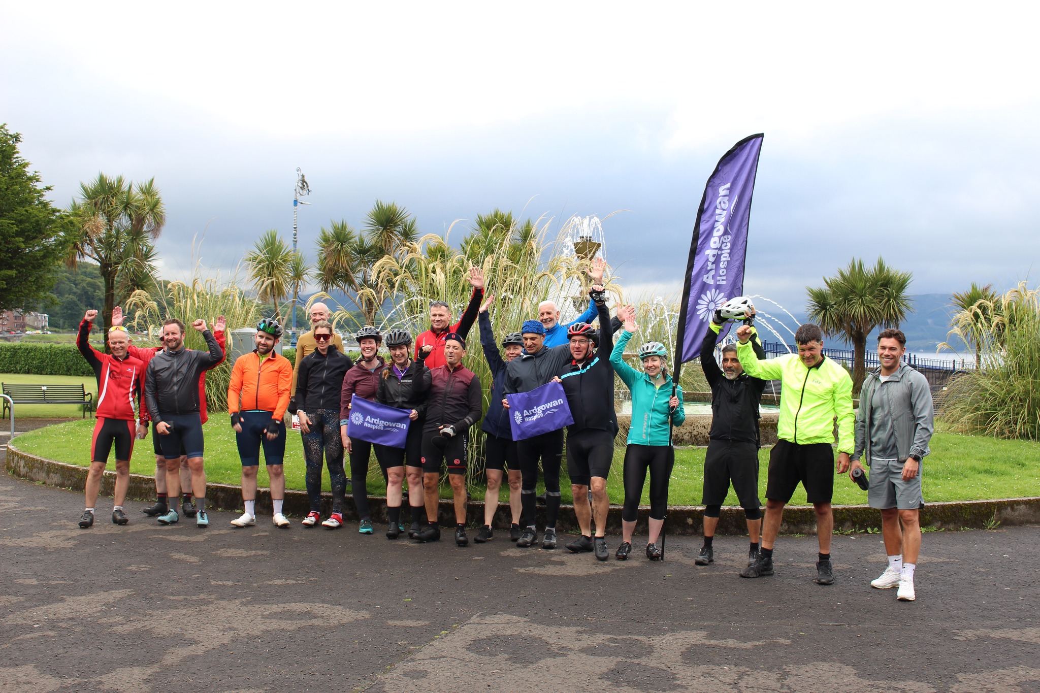 A large group of cyclists pose and celebrate outdoors near a fountain and palm trees. Many wear bright cycling gear and helmets. Several people hold "Ardgowan Hospice" flags, and a tall hospice-branded banner is visible. The group appears cheerful, raising arms in triumph, likely after completing a charity ride.